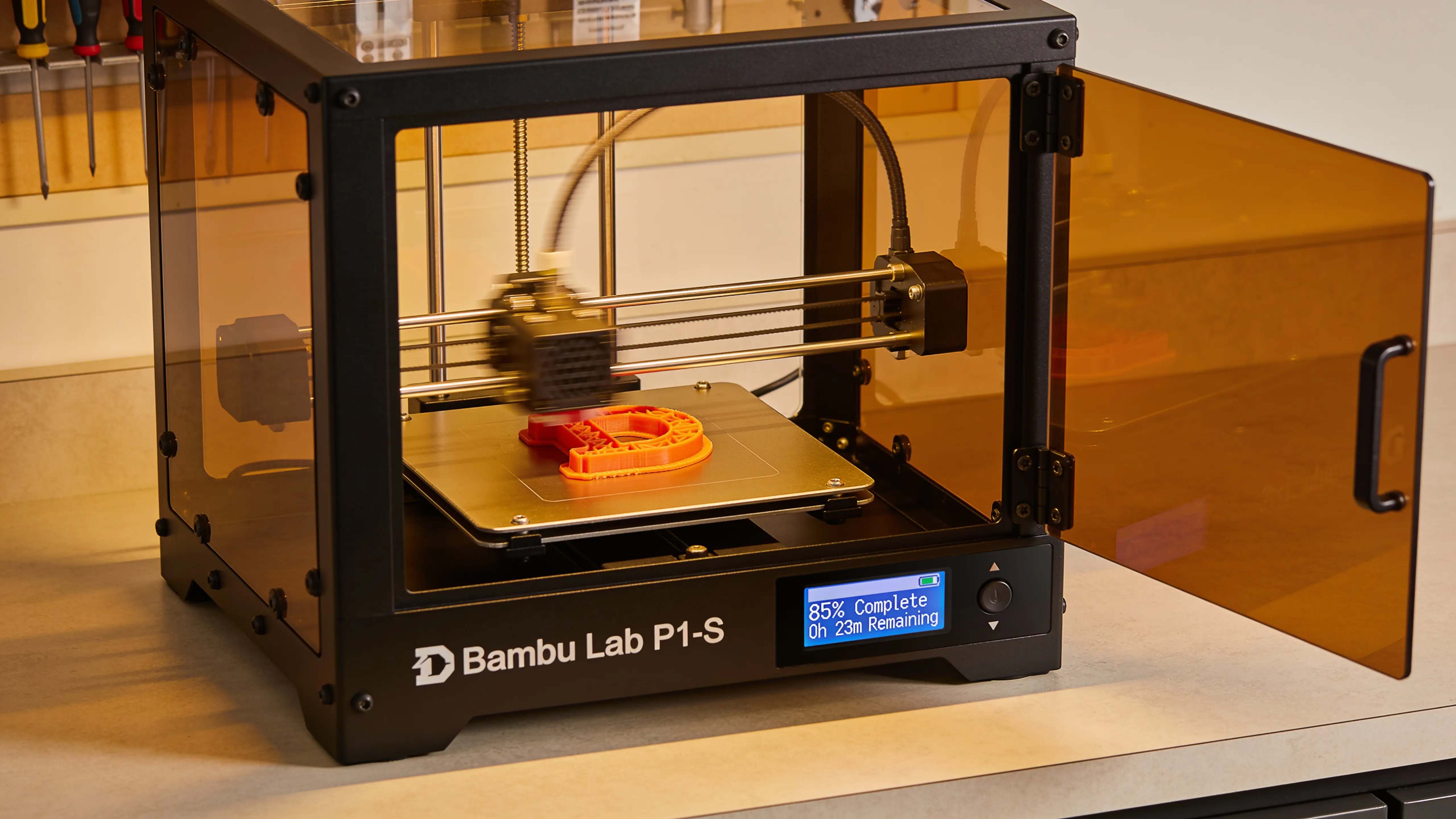 Enclosed Bambu Lab P1S 3D printer mid-print with an orange PLA part visible and the LCD screen showing print progress and time remaining